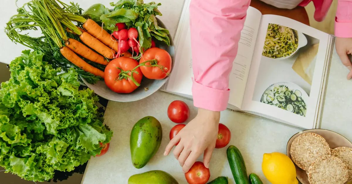 Mutfak tezgâhında taze sebzeler ve meyveler; havuç, turp, domates, marul, avokado, salatalık ve limonla birlikte açık bir yemek kitabı